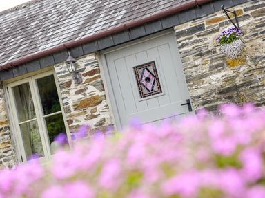 A stone cottage exterior with a grey door featuring stained glass and a window with curtains near a hanging flower basket at Mimi's Cottage in St Neot