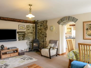 A living room with an armchair television and wood burning stove next to a dining area at Mimis Cottage in St Neot