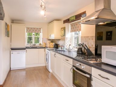 A kitchen with white cabinets a gas stove a washing machine a dishwasher and windows with patterned blinds at Mimi's Cottage in St Neot