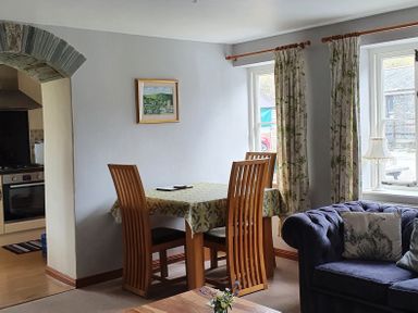 A dining area with a wooden table and four chairs beside windows with curtains and a sofa with cushions at Mimi's Cottage in St Neot