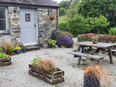 An outdoor patio area with a wooden picnic table and benches surrounded by potted plants and a stone building with a painted door at Mimi's Cottage in St Neot