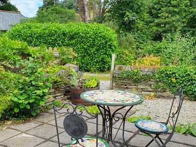 An outdoor patio with a round mosaic table and two chairs surrounded by greenery and stone walls at Mimi's Cottage in St Neot