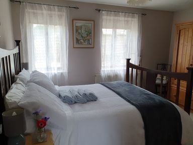 A bedroom with a double bed with white bedding and folded towels two windows with sheer curtains a wooden wardrobe and a small bedside table with a lamp and flowers at Mimis Cottage in St Neot