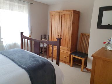 A bedroom with a wooden bed frame a wooden wardrobe two chairs a wooden dresser with flowers and a mirror at Mimis Cottage in St Neot
