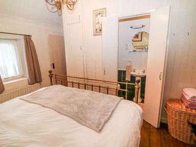 A bedroom with a metal bed frame a window with curtains an open door showing a sink and mirror and a wicker basket with folded towels at Mary Rose Cottage in Cordal near Castleisland County Kerry