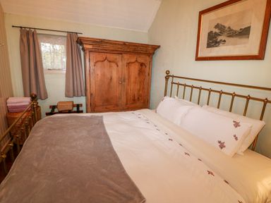 A bedroom with a bed with white and floral bedding a wooden wardrobe a window with curtains and a framed picture on the wall at Mary Rose Cottage in Cordal near Castleisland County Kerry