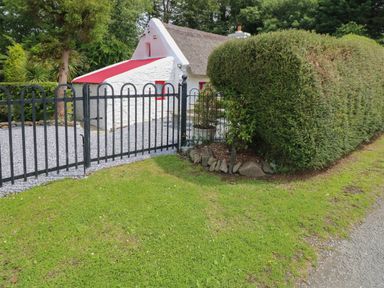 A white stone cottage with a thatched roof behind a black metal gate and trimmed hedges at Mary Rose Cottage in Cordal near Castleisland County Kerry