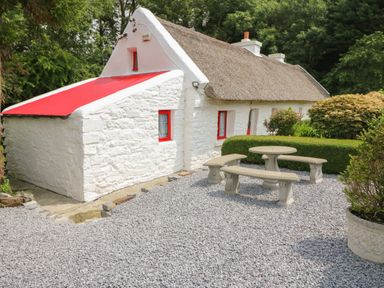 A white stone cottage with red window frames and a thatched roof surrounded by trees and bushes with a gravel patio and stone table and benches at Mary Rose Cottage in Cordal near Castleisland County Kerry