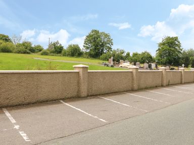 A parking lot with marked spaces next to a stone wall and a cemetery behind the wall at Mary Rose Cottage in Cordal near Castleisland County Kerry