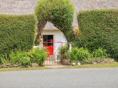 A red metal gate with an archway of trimmed greenery leading to a white building with a thatched roof at Mary Rose Cottage in Cordal near Castleisland County Kerry