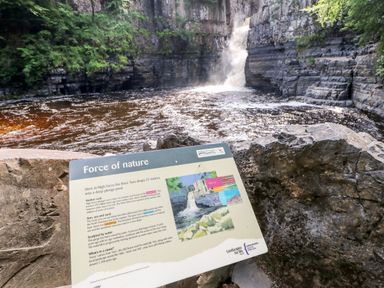 A waterfall cascading into a river surrounded by rock cliffs with an informational sign about the force of nature at Badgers Sett Winston near Staindrop