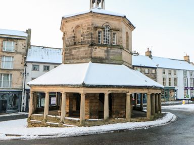 A stone octagonal market cross with pillars and a snow covered roof in a town square with surrounding buildings at Badgers Sett in Winston near Staindrop