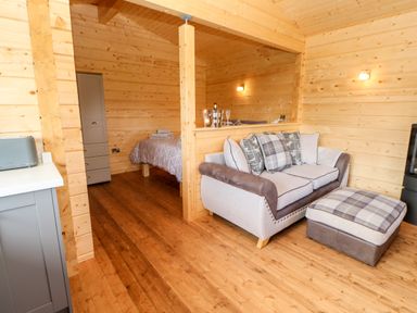 A wooden interior with a beige and brown sofa with plaid cushions and an ottoman next to a partially visible kitchen and a small bedroom area with a bed at Badgers Sett in Winston near Staindrop