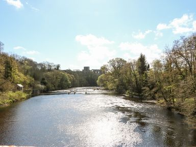 A river flowing through a wooded area with a castle ruin in the background at Badgers Sett in Winston near Staindrop