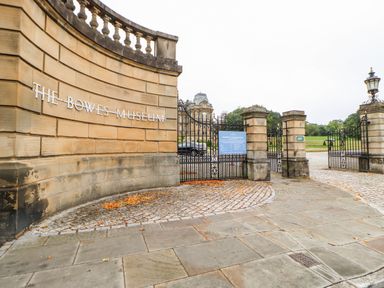 The entrance gate and stone wall with sign for The Bowes Museum and a building in the background at Badgers Sett in Winston near Staindrop