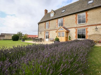 A stone house with a wooden porch and a patio with outdoor furniture next to a garden with purple flowers at Shifford Manor Farm in Shifford near Bampton Oxfordshire