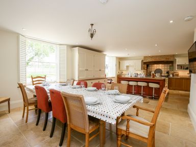 A dining area with a large table set with plates and glasses surrounded by various chairs next to a kitchen with stools at the counter and built in cupboards at Shifford Manor Farm in Shifford near Bampton Oxfordshire