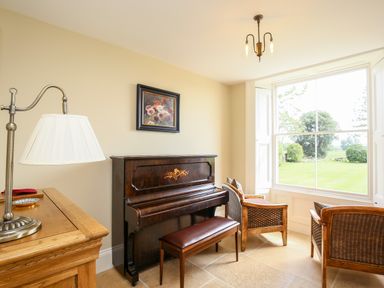 A room with a wooden piano and bench next to two wicker chairs by a large window at Shifford Manor Farm in Shifford near Bampton Oxfordshire