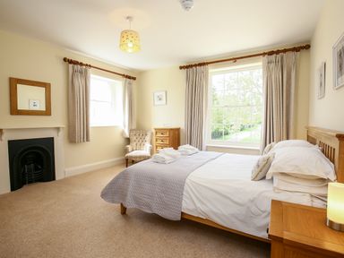 A bedroom with a bed a chair a wooden chest of drawers and windows with curtains at Shifford Manor Farm in Shifford near Bampton Oxfordshire