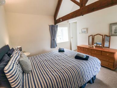 A bedroom with a striped bedspread two rolled towels on the bed a wooden dresser with a three-panel mirror and a window with curtains at Silver Birches in Glaisdale