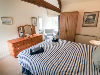 A bedroom with a striped bedspread two rolled towels on the bed a wooden dresser with mirror a wooden wardrobe an armchair and framed pictures at Silver Birches in Glaisdale