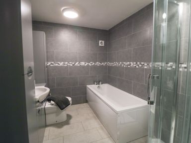 A bathroom with a bathtub shower cabin toilet and sink with grey tiled walls and light floor tiles at Silver Birches in Glaisdale