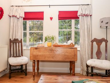 A dining area with a wooden table between two wooden chairs with cushions and red window blinds at October Cottage in Chalford Gloucestershire