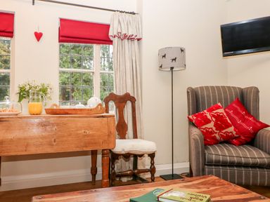 A room with a wooden table and chair near a window a striped armchair with red cushions a floor lamp and a wall-mounted television at October Cottage in Chalford Gloucestershire