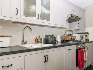A kitchen with white cabinets a sink a toaster kettle stove with a red towel and a basket on the counter at October Cottage in Chalford Gloucestershire
