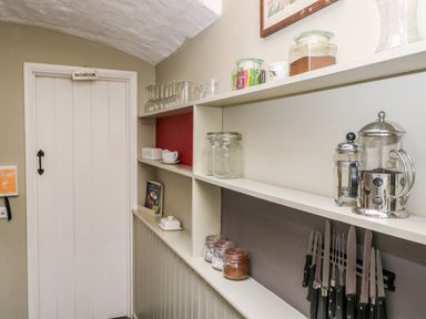 A hallway with white shelves holding jars, glasses, coffee makers, and knives next to a door labeled bathroom at October Cottage in Chalford Gloucestershire