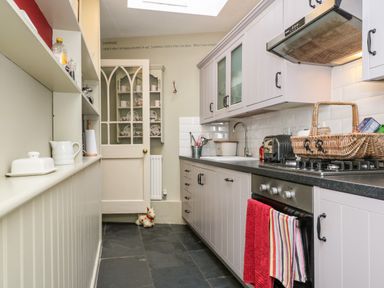 A galley kitchen with white cabinets and black countertop next to a glass panel door with dishes on shelves at October Cottage in Chalford Gloucestershire