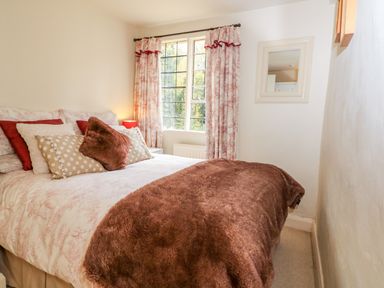 A bedroom with a bed covered with multiple pillows and a brown blanket next to a window with curtains at October Cottage in Chalford Gloucestershire