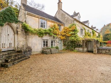 A stone cottage with climbing plants on the walls a gravel driveway and wooden doors at October Cottage in Chalford Gloucestershire