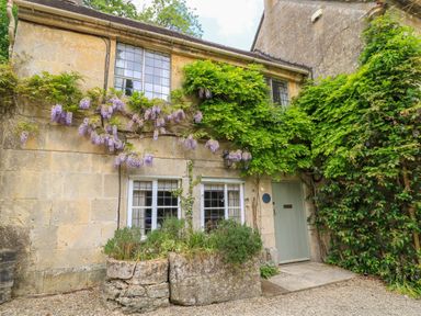 Stone cottage exterior with climbing plants and flowers near a green door at October Cottage in Chalford Gloucestershire