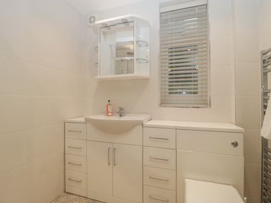 A bathroom with a white sink cabinet, mirror with shelves, window blinds, and towel rack at Greenlawns in Burnham-On-Sea