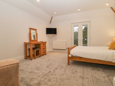 A bedroom with a wooden bed a dressing table with mirror a black stool a wall-mounted television and a set of French doors at Greenlawns in Burnham-On-Sea