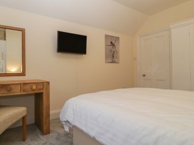 A bedroom with a bed a wooden dressing table a chair a wall-mounted tv and a painting at Greenlawns in Burnham-On-Sea