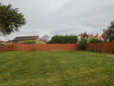 A grassy backyard enclosed by wooden fences with trees and houses in the background at Greenlawns in Burnham-On-Sea