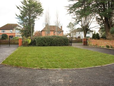 A front yard with a lawn surrounded by a driveway and two gates with houses and trees in the background at Greenlawns in Burnham-On-Sea