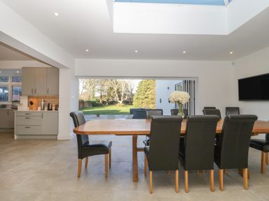 A dining area with a wooden table and eight black chairs next to an open patio door showing a garden at Greenlawns in Burnham-On-Sea