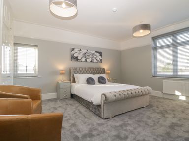 A bedroom with a bed with tufted headboard and footboard two bedside tables with lamps two brown armchairs and two windows at Greenlawns in Burnham-On-Sea