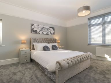 A bedroom with a padded bed a gray carpet two nightstands with lamps and a window with blinds at Greenlawns in Burnham-On-Sea