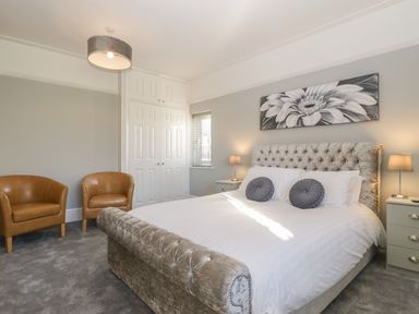 A bedroom with a bed two round pillows two bedside tables with lamps two brown armchairs and a floral painting above the bed at Greenlawns in Burnham-On-Sea