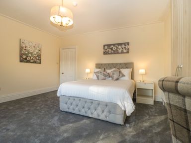 A bedroom with a bed two bedside tables with lamps paintings on walls and a checkered armchair at Greenlawns in Burnham-On-Sea