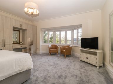 A bedroom with two brown chairs in a bay window a bed a television on a stand and built in wardrobe with a vanity at Greenlawns in Burnham-On-Sea