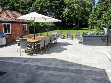 An outdoor patio with a long wooden dining table and chairs under umbrellas a barbecue grill lounge chairs on grass and a hot tub at Greenlawns in Burnham-On-Sea