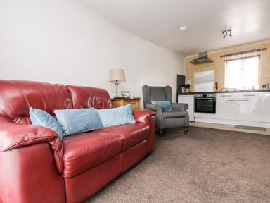 A living room with a red leather sofa blue cushions a grey armchair a wooden side table with a lamp and a kitchen area at Coach House Cottage in Bude