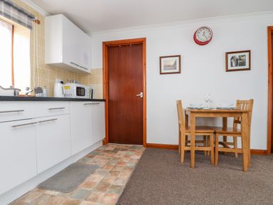 A kitchen area with white cabinets microwave and kettle next to a wooden dining table with two chairs at Coach House Cottage in Bude