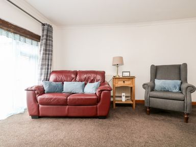 A red leather sofa with blue cushions a wooden side table with a lamp and picture frame a grey armchair with a blue cushion and a window with curtains at Coach House Cottage in Bude