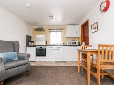 A kitchen with white cabinets a small dining table with chairs and a gray armchair at Coach House Cottage in Bude